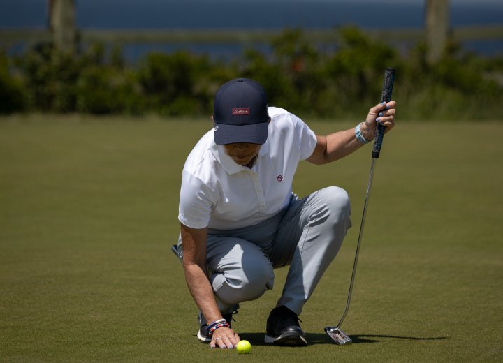 Golfer crouches on the putting green, holding a putter while carefully reading the line before a putt on a sunny day.