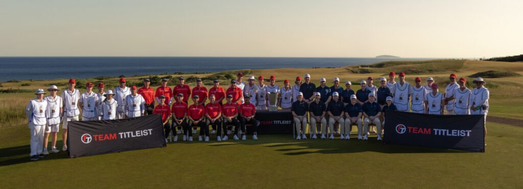 Large group of golfers, caddies, and staff pose on a coastal golf course behind “Team Titleist” banners, with the ocean and rolling hills visible in the background.