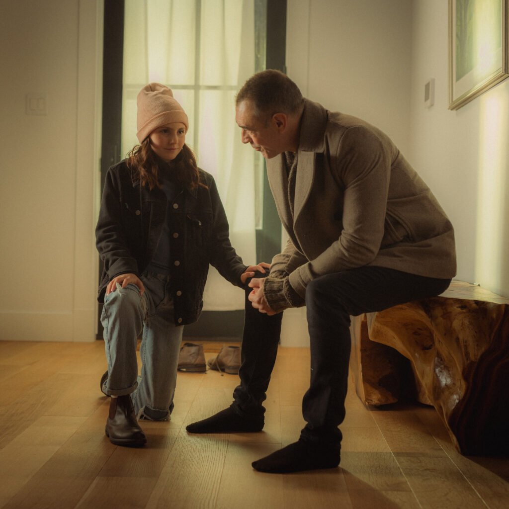 A man crouches to speak gently with a young child wearing a pink knit hat and coat, inside a softly lit home entryway